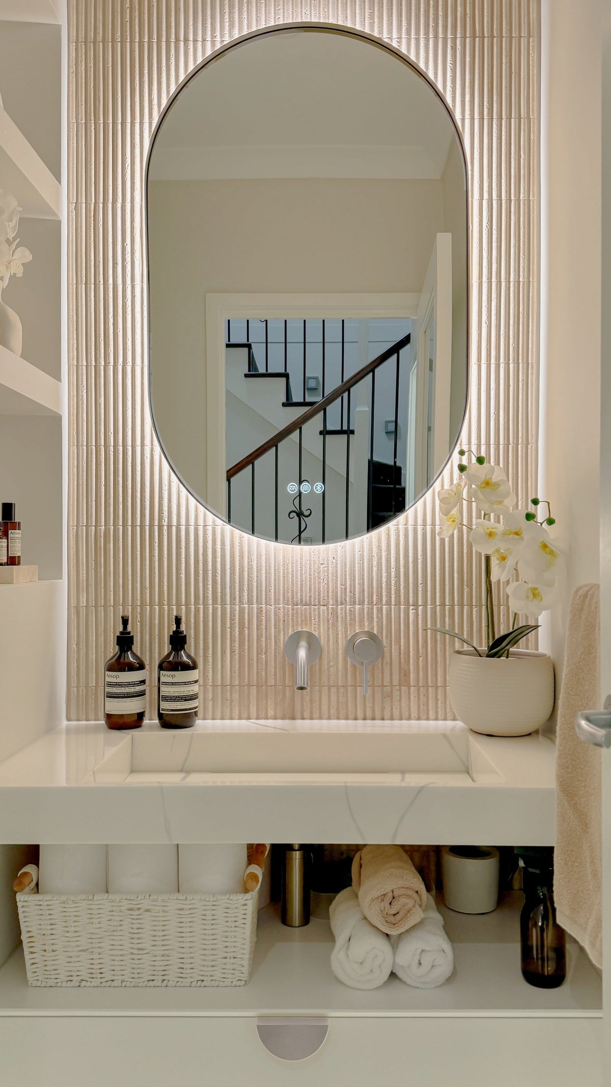 The Valencia 600 x 1000 mm oval framed backlit LED mirror in Brushed Nickel, above a white stone basin with a matching Milano wall spout and mixer, fluted travertine wall