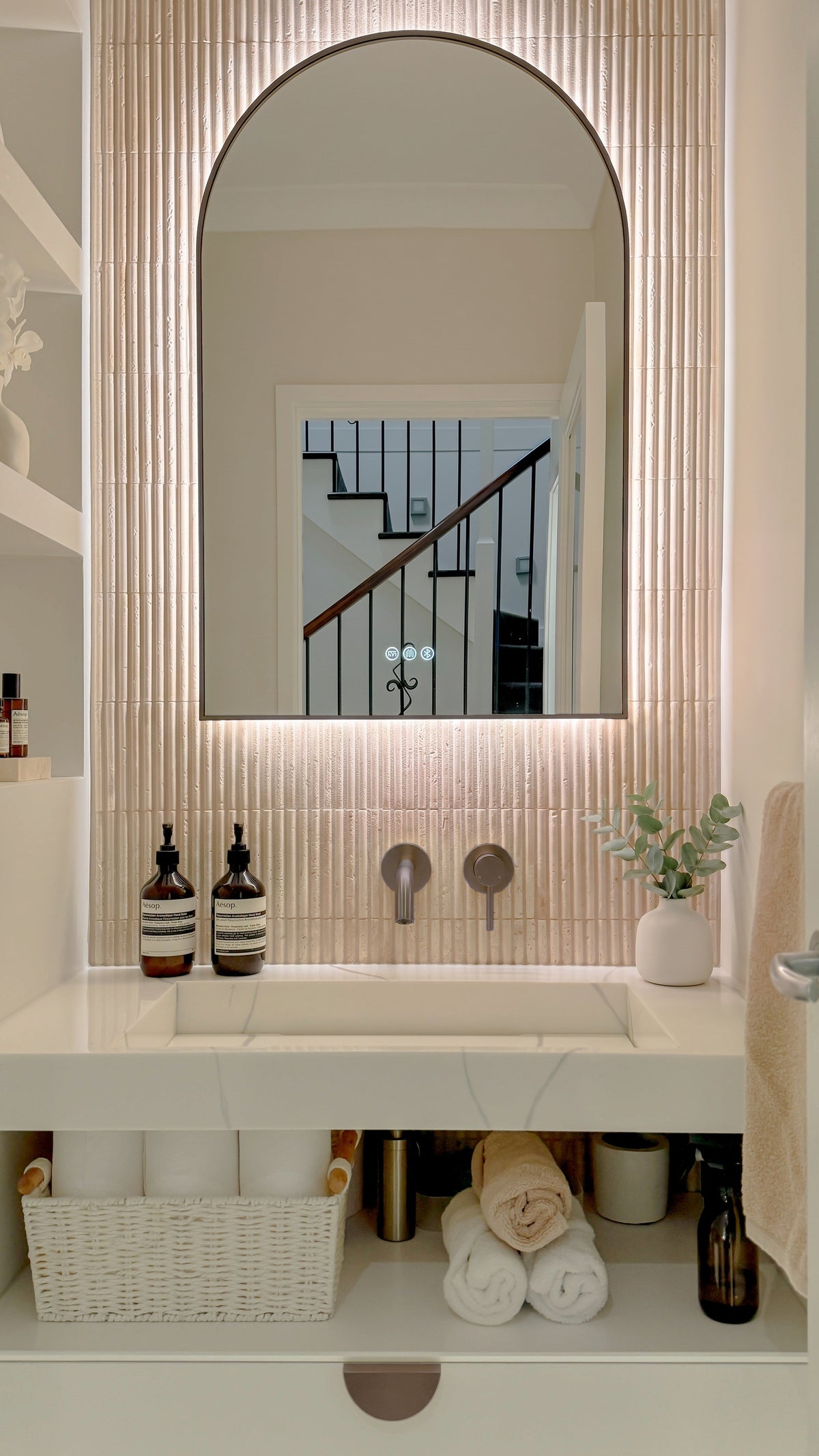 The Marbella 600 x 900 mm arch framed backlit LED mirror in Antique Brass, above a white stone basin with a matching Milano wall spout and mixer, fluted travertine wall