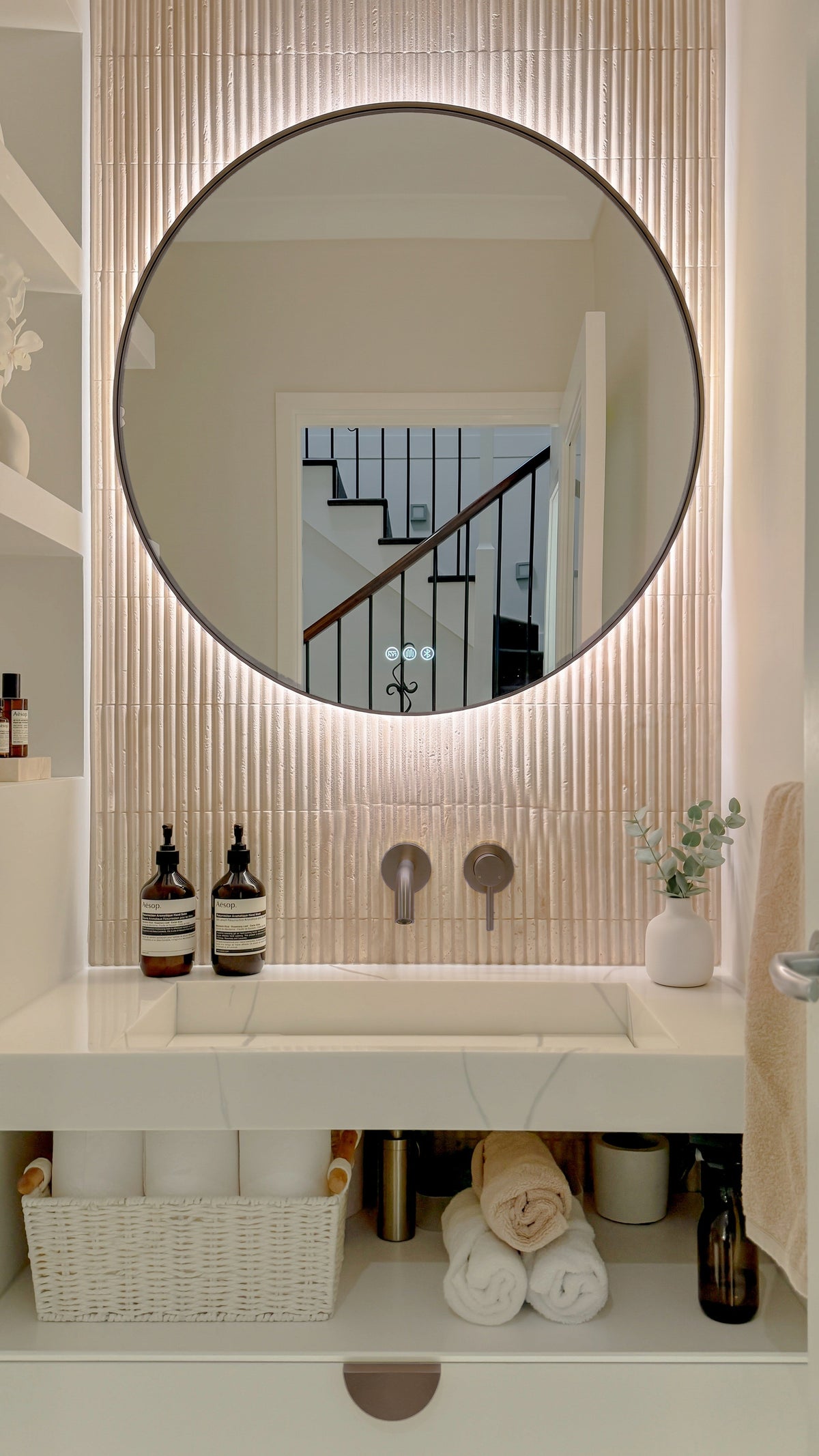 The Capri round 800 mm framed backlit LED mirror in Antique Brass, above a white stone basin with a matching Milano wall spout and mixer, fluted travertine wall