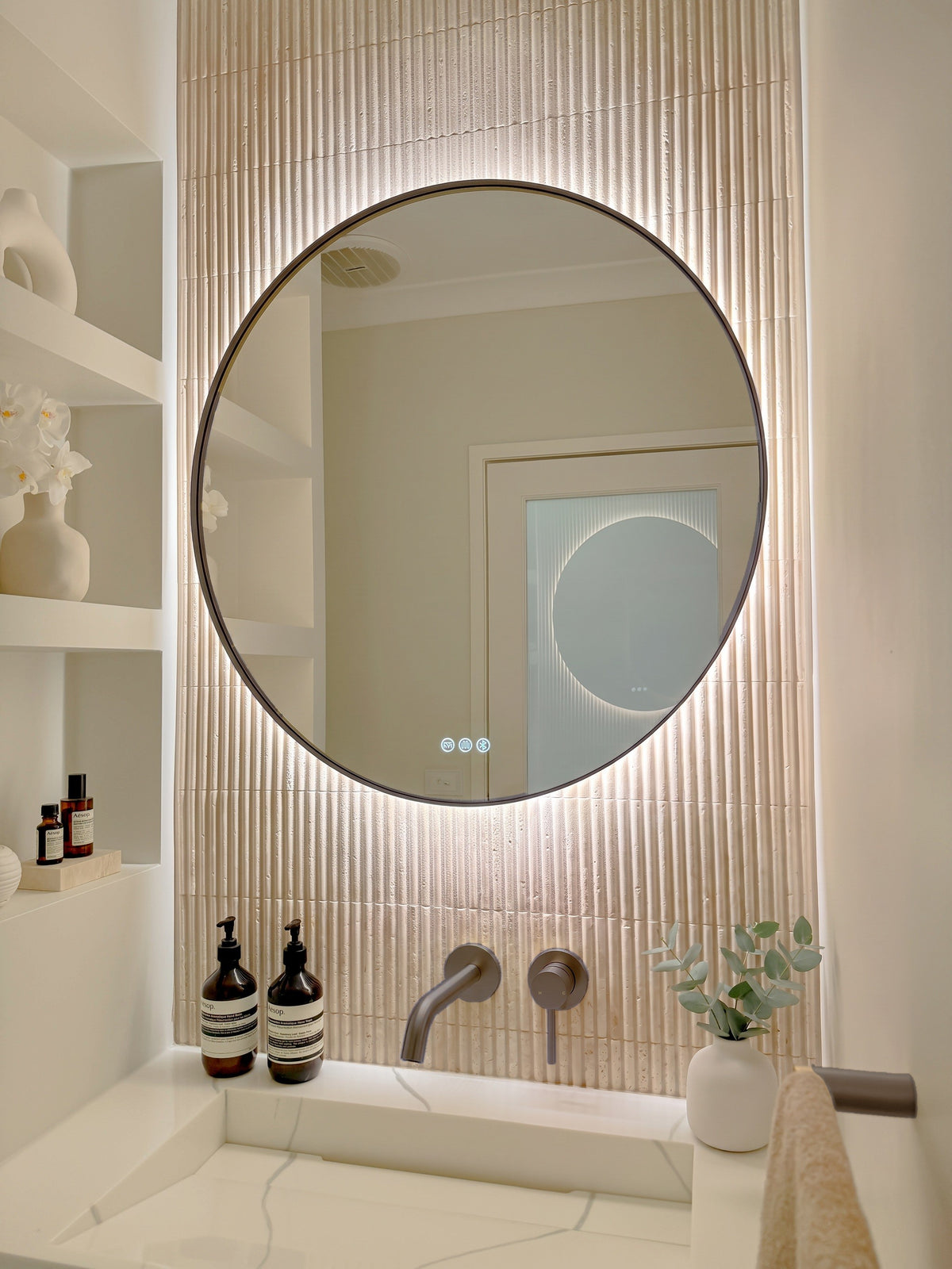 The Capri round 800 mm framed backlit LED mirror in Antique Brass, angled close view with the warm backlight glowing on fluted travertine
