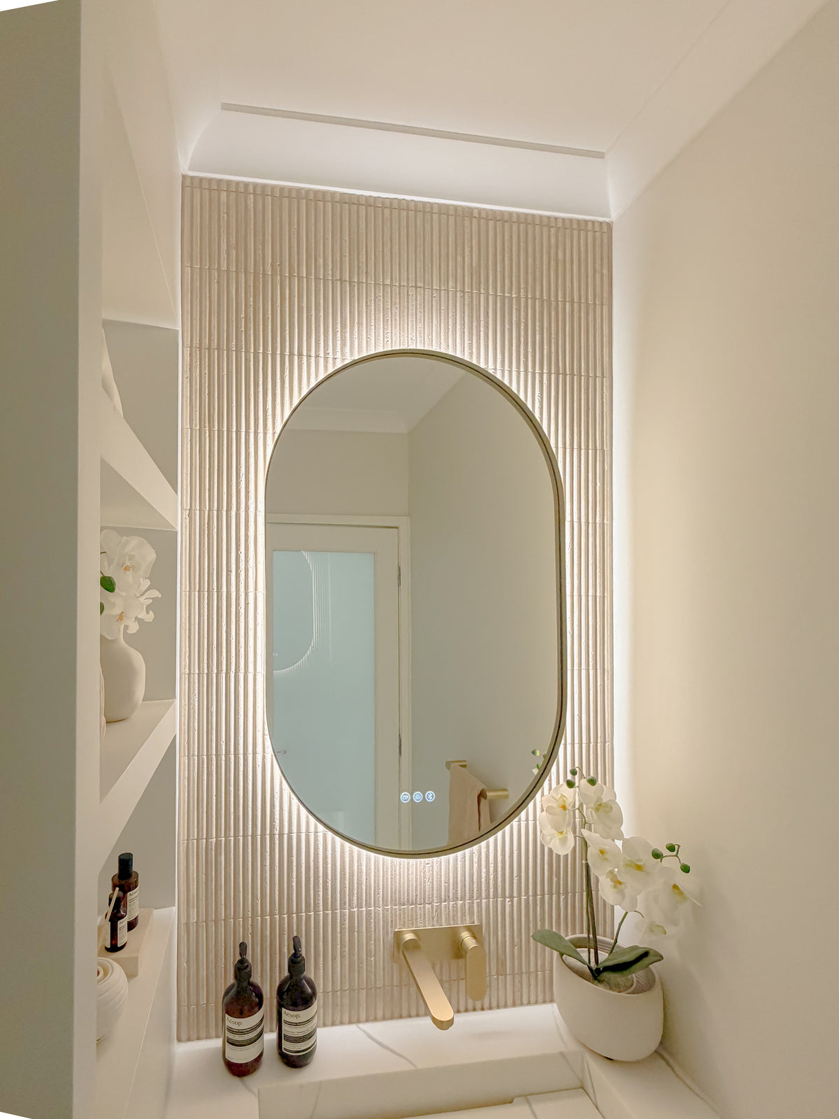 The Valencia oval mirror lit warm against fluted tiles, orchid beside the basin