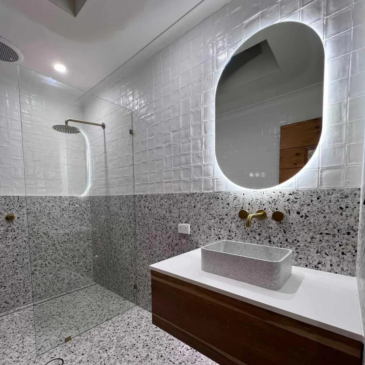 Backlit vertical oval LED mirror above a terrazzo bathroom vanity with a brass wall mixer