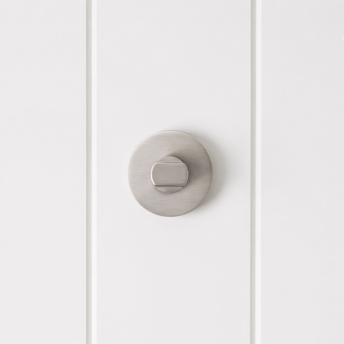 Prague Door Lock in Brushed Nickel, front view of the thumb-turn on its round rose, white vertical-panelled wall
