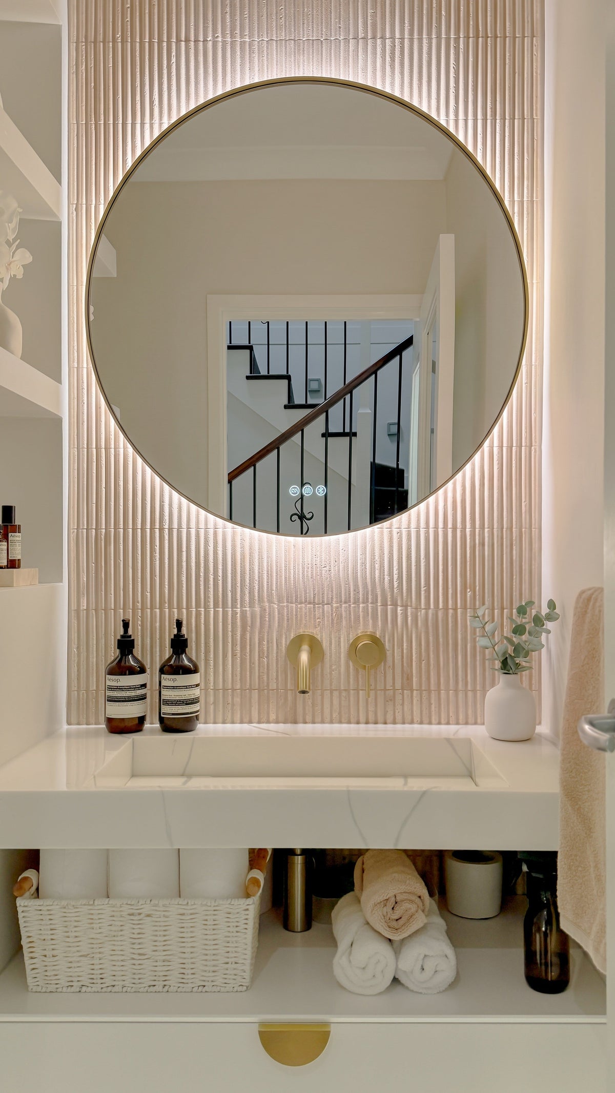The Capri round 800 mm framed backlit LED mirror in Brushed Brass, above a white stone basin with a matching Milano wall spout and mixer, fluted travertine wall