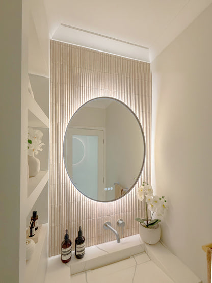 The Capri round 800 mm framed backlit LED mirror in Brushed Nickel, seen from the side on a backlit fluted travertine wall, showing the slim frame edge