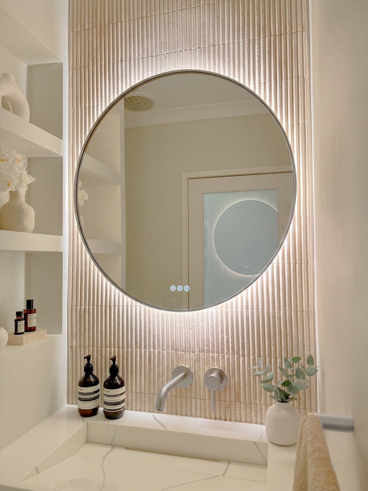 The Capri round 800 mm framed backlit LED mirror in Brushed Nickel, angled close view with the warm backlight glowing on fluted travertine