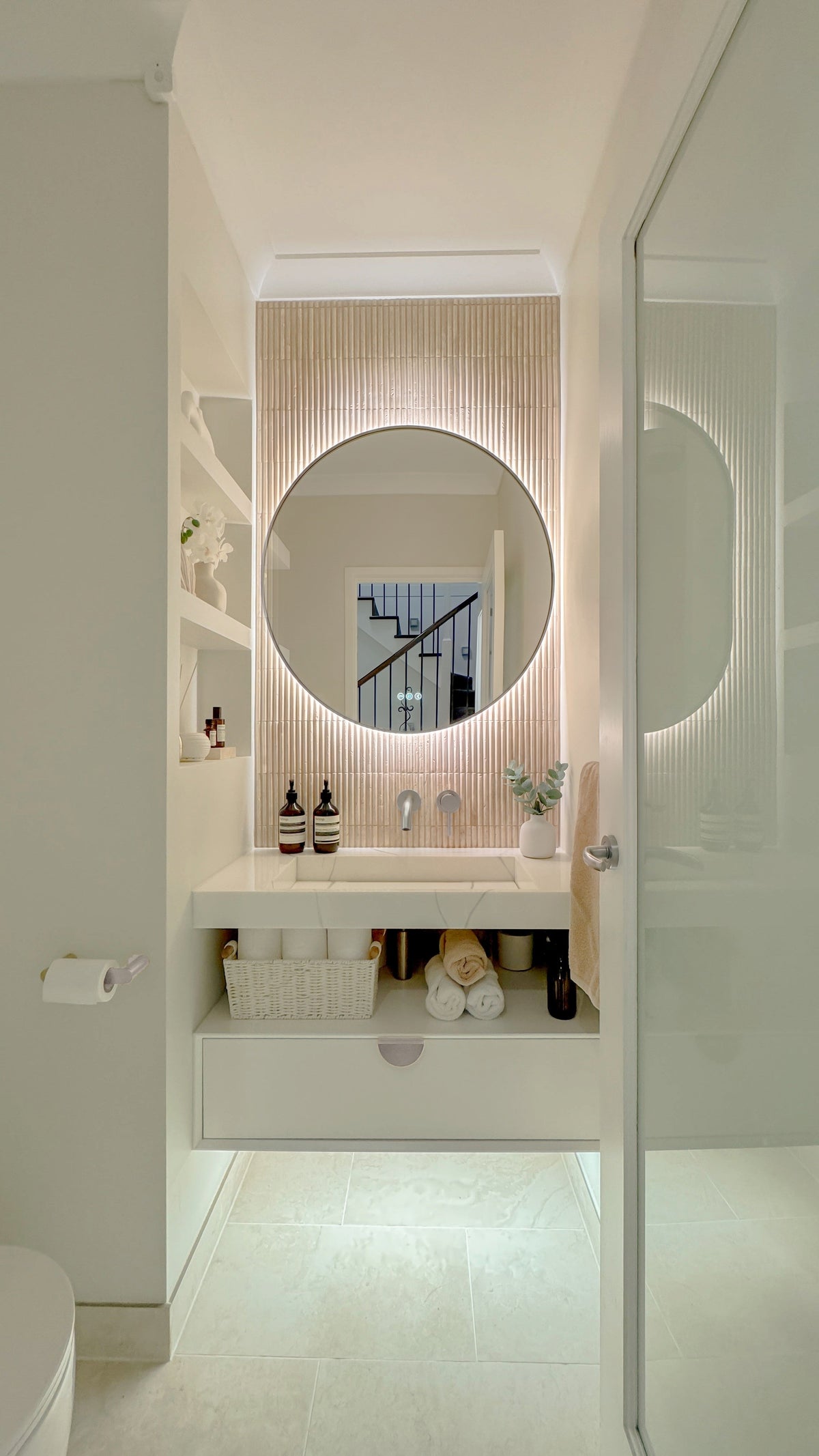 The Capri round 800 mm framed backlit LED mirror in Brushed Nickel, seen through the doorway of a calm white powder room