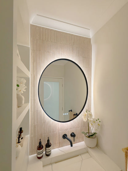 The Capri round 800 mm framed backlit LED mirror in Matte Black, seen from the side on a backlit fluted travertine wall, showing the slim frame edge