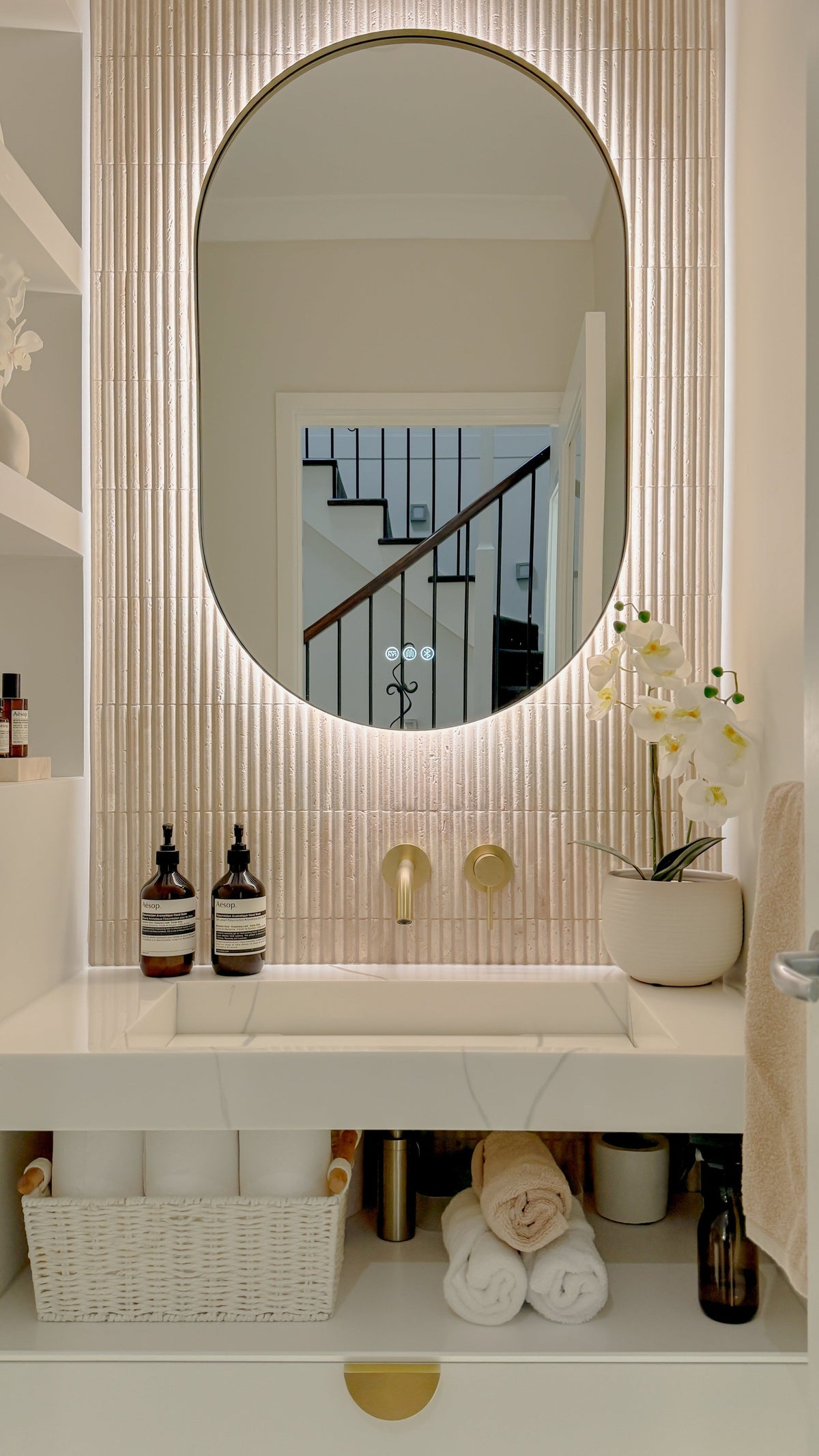 The Valencia 600 x 1000 mm oval framed backlit LED mirror in Brushed Brass, above a white stone basin with a matching Milano wall spout and mixer, fluted travertine wall