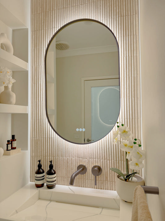 The Valencia 600 x 900 mm oval framed backlit LED mirror in Antique Brass, angled close view with the warm backlight glowing on fluted travertine