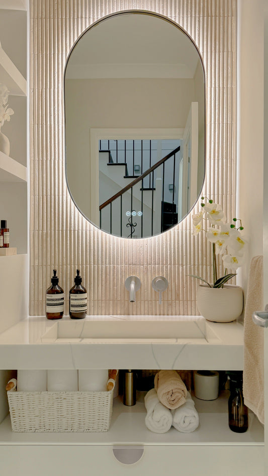 The Valencia 600 x 900 mm oval framed backlit LED mirror in Brushed Nickel, above a white stone basin with a matching Milano wall spout and mixer, fluted travertine wall