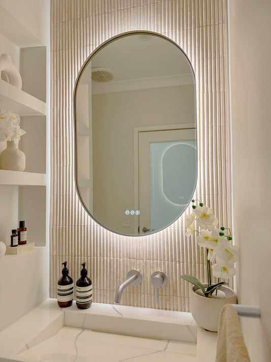 The Valencia 600 x 900 mm oval framed backlit LED mirror in Brushed Nickel, angled close view with the warm backlight glowing on fluted travertine
