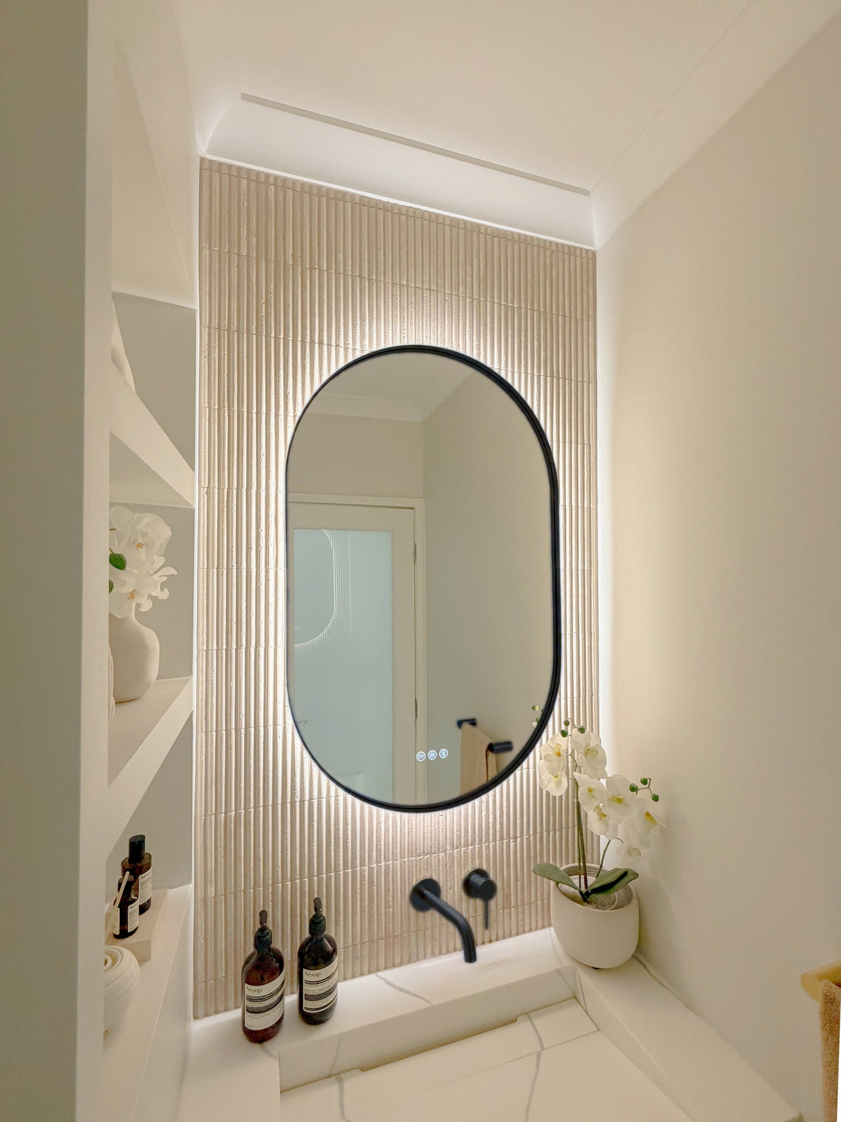 The Valencia 600 x 1000 mm oval framed backlit LED mirror in Matte Black, seen from the side on a backlit fluted travertine wall, showing the slim frame edge