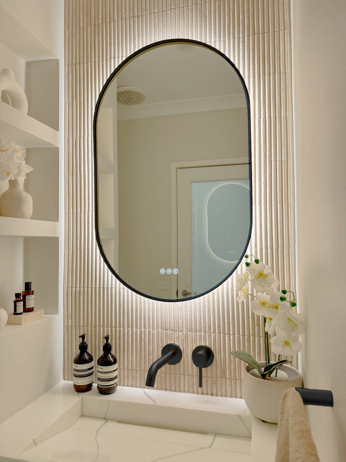 The Valencia 600 x 1000 mm oval framed backlit LED mirror in Matte Black, angled close view with the warm backlight glowing on fluted travertine