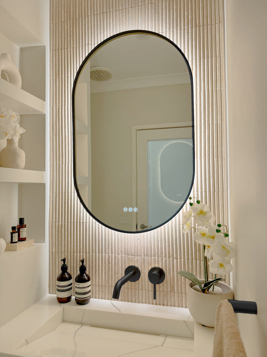 The Valencia 600 x 900 mm oval framed backlit LED mirror in Matte Black, angled close view with the warm backlight glowing on fluted travertine