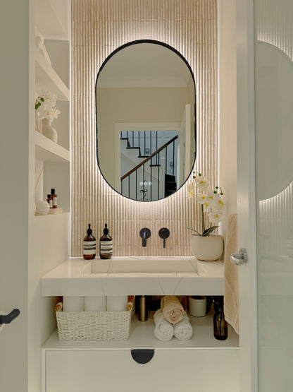 The Valencia 600 x 1000 mm oval framed backlit LED mirror in Matte Black, in a powder room above a floating white vanity with matching hardware