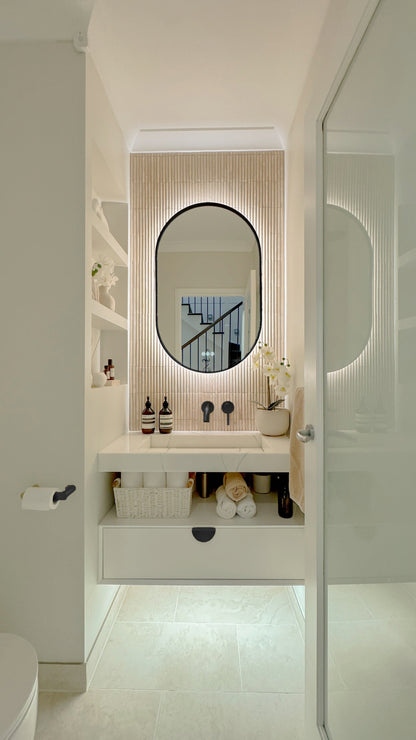 The Valencia 600 x 1000 mm oval framed backlit LED mirror in Matte Black, seen through the doorway of a calm white powder room
