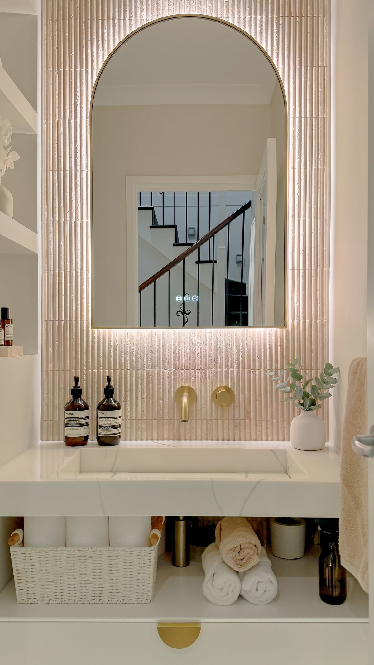 The Marbella 600 x 900 mm arch framed backlit LED mirror in Brushed Brass, above a white stone basin with a matching Milano wall spout and mixer, fluted travertine wall