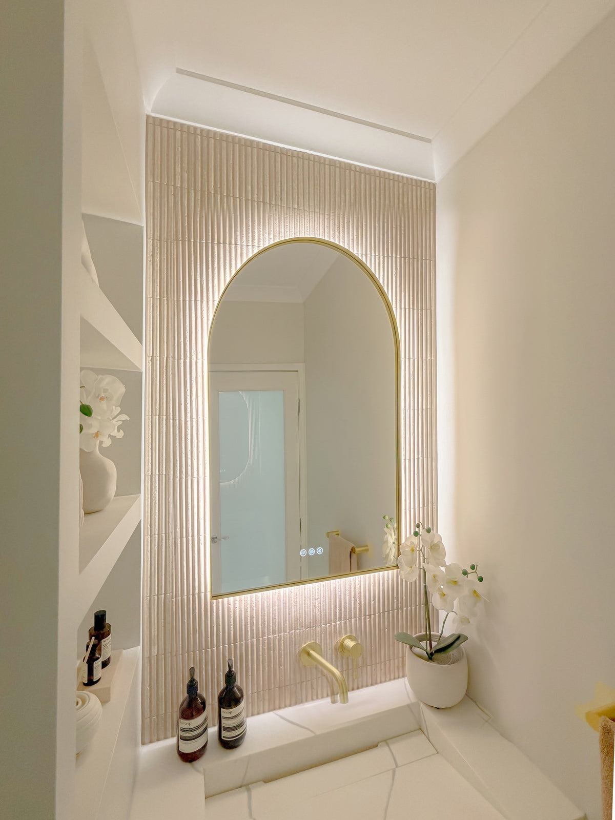 The Marbella 600 x 900 mm arch framed backlit LED mirror in Brushed Brass, seen from the side on a backlit fluted travertine wall, showing the slim frame edge