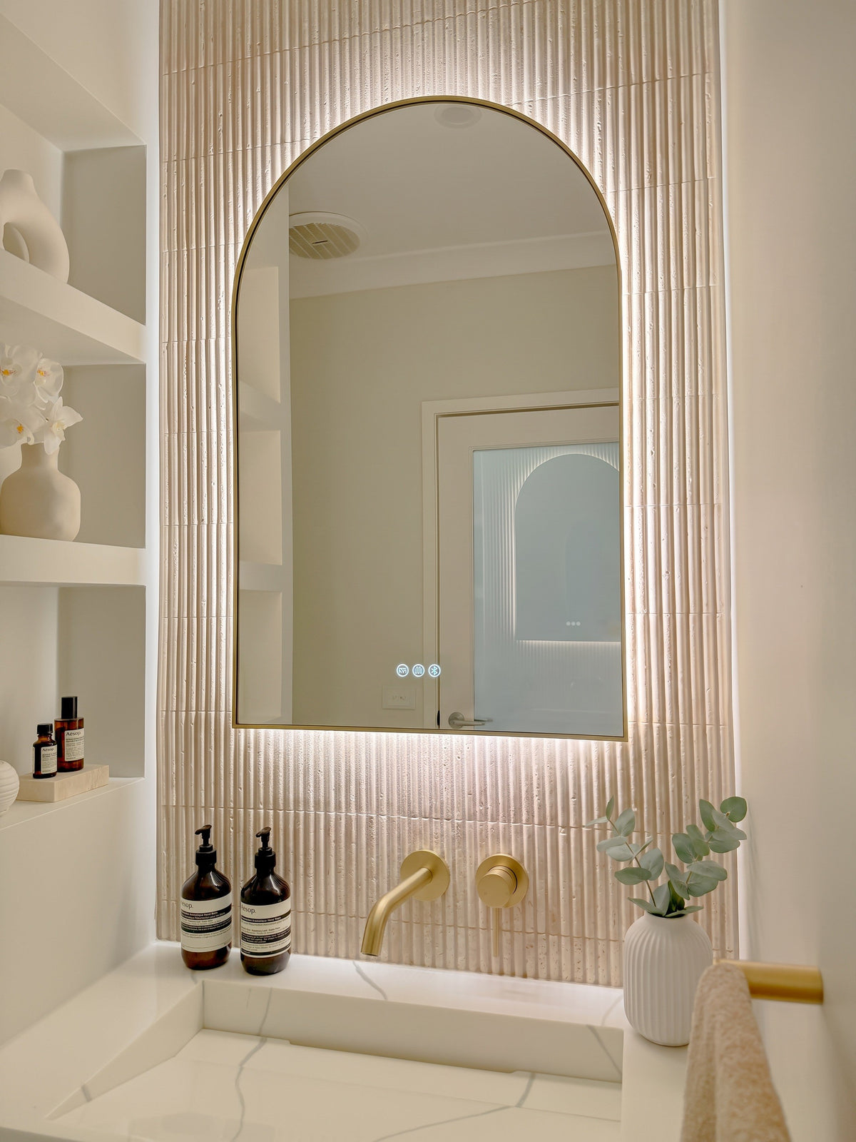 The Marbella 600 x 900 mm arch framed backlit LED mirror in Brushed Brass, angled close view with the warm backlight glowing on fluted travertine