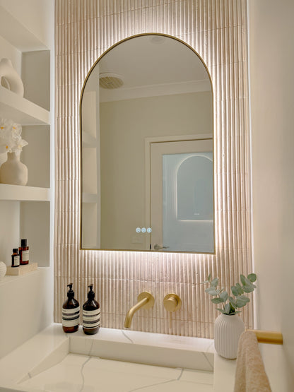 The Marbella 600 x 900 mm arch framed backlit LED mirror in Brushed Brass, angled close view with the warm backlight glowing on fluted travertine