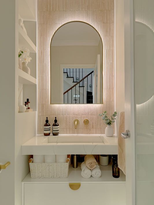 The Marbella 600 x 900 mm arch framed backlit LED mirror in Brushed Brass, in a powder room above a floating white vanity with matching hardware