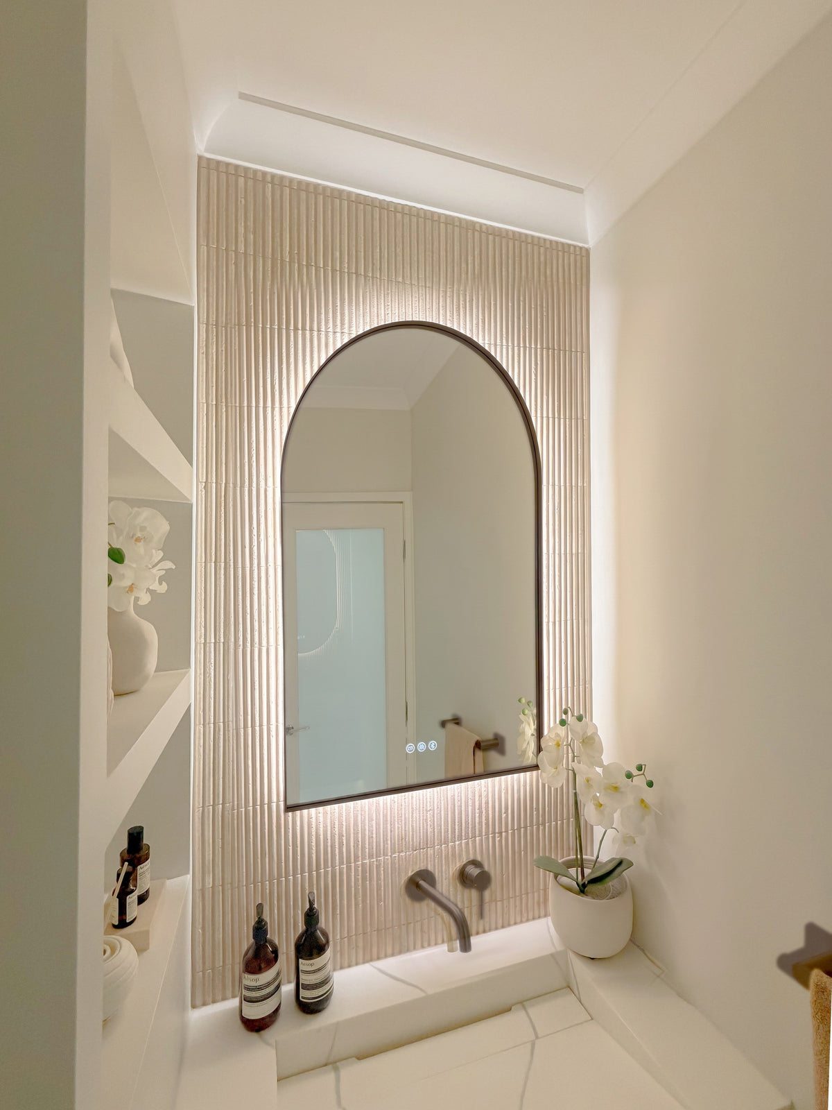 The Marbella 600 x 900 mm arch framed backlit LED mirror in Antique Brass, seen from the side on a backlit fluted travertine wall, showing the slim frame edge