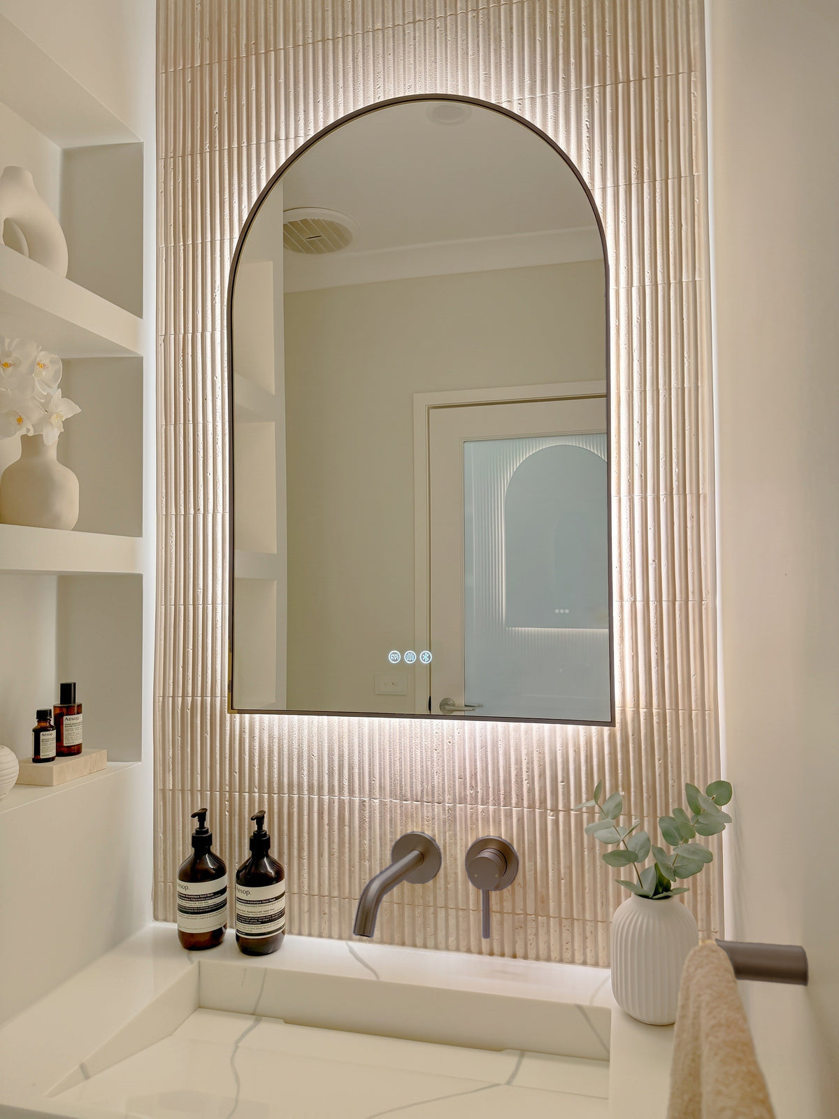 The Marbella 600 x 900 mm arch framed backlit LED mirror in Antique Brass, angled close view with the warm backlight glowing on fluted travertine