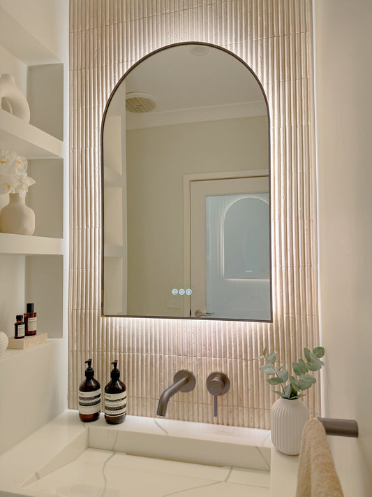 The Marbella 600 x 1000 mm arch framed backlit LED mirror in Antique Brass, angled close view with the warm backlight glowing on fluted travertine