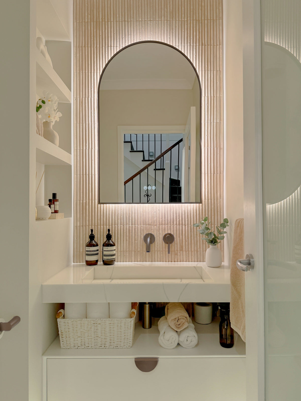 The Marbella 600 x 900 mm arch framed backlit LED mirror in Antique Brass, in a powder room above a floating white vanity with matching hardware