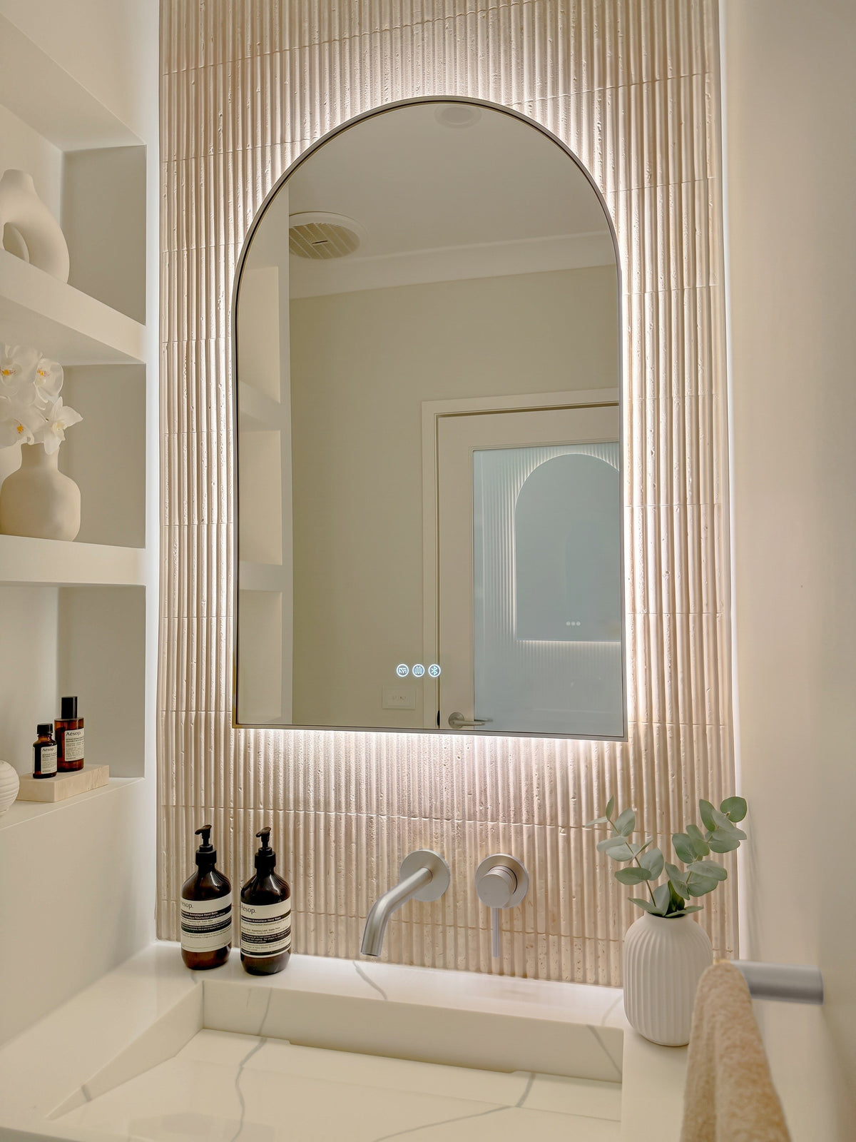 The Marbella 600 x 900 mm arch framed backlit LED mirror in Brushed Nickel, angled close view with the warm backlight glowing on fluted travertine