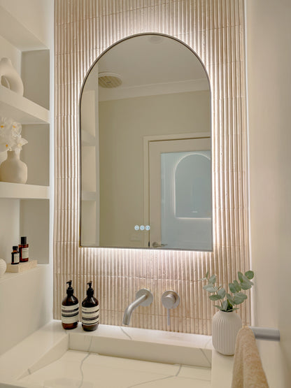 The Marbella 600 x 900 mm arch framed backlit LED mirror in Brushed Nickel, angled close view with the warm backlight glowing on fluted travertine