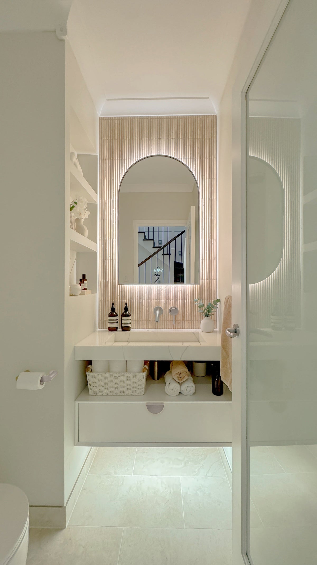 The Marbella 600 x 900 mm arch framed backlit LED mirror in Brushed Nickel, seen through the doorway of a calm white powder room