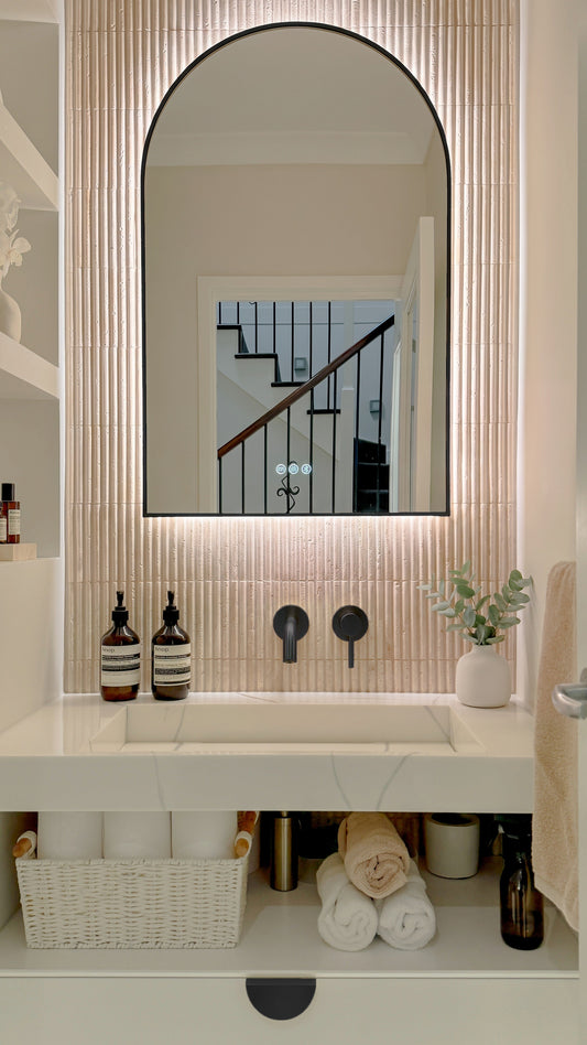 The Marbella 600 x 900 mm arch framed backlit LED mirror in Matte Black, above a white stone basin with a matching Milano wall spout and mixer, fluted travertine wall