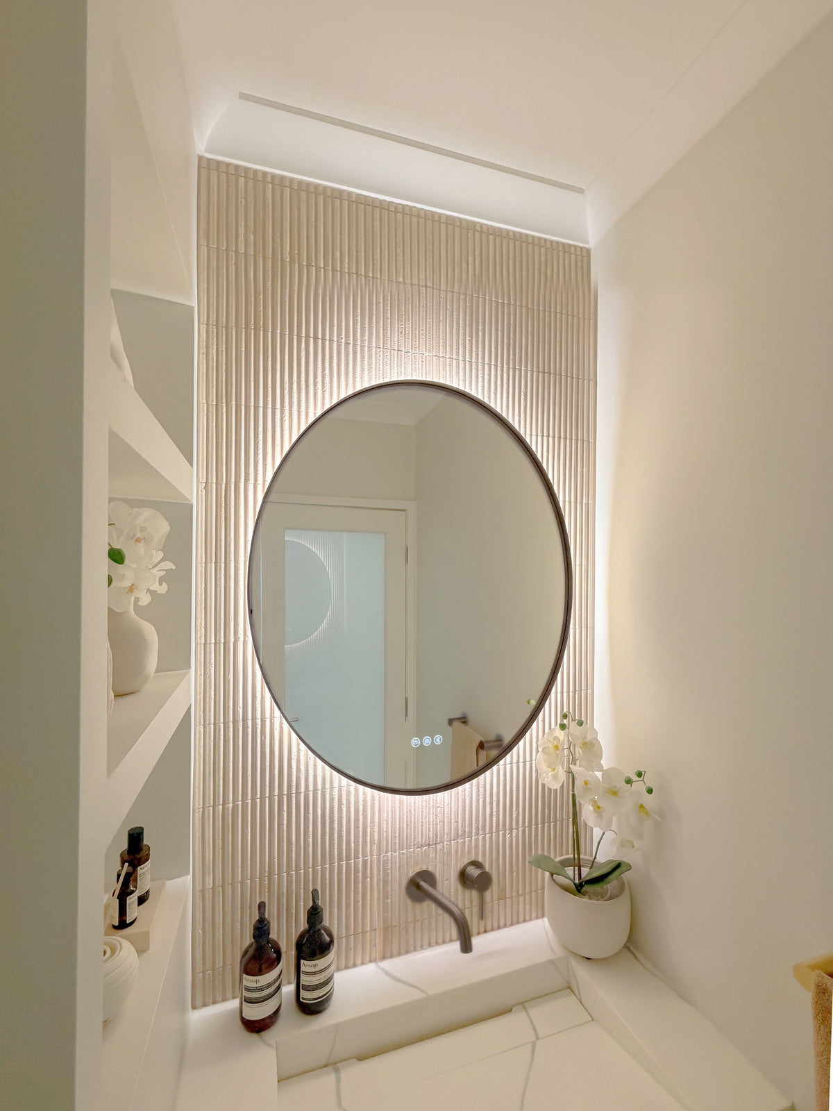 The Capri round 800 mm framed backlit LED mirror in Antique Brass, seen from the side on a backlit fluted travertine wall, showing the slim frame edge