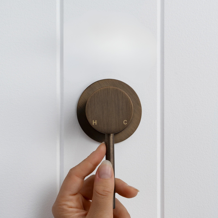 Milano Wall Mixer in Antique Brass, hand on the pin lever, front view on a white vertical-panelled wall