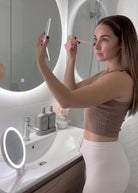 Woman using foldable LED Mini Make-up mirror in front of big Circle LED mirror in white bathroom