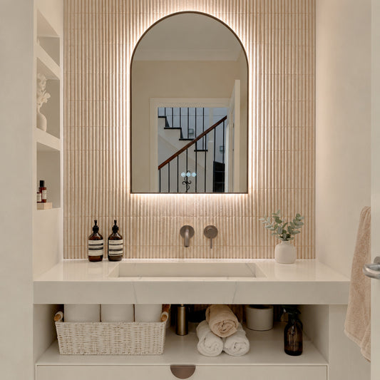 The Marbella 600 x 1000 mm arch framed backlit LED mirror in Antique Brass, lit and seen straight-on above a white stone basin with a matching wall spout and mixer