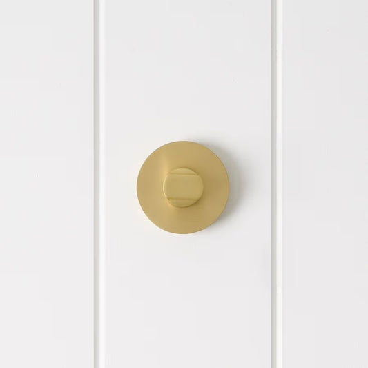 Prague Door Lock in Brushed Brass, front view of the thumb-turn on its round rose, white vertical-panelled wall