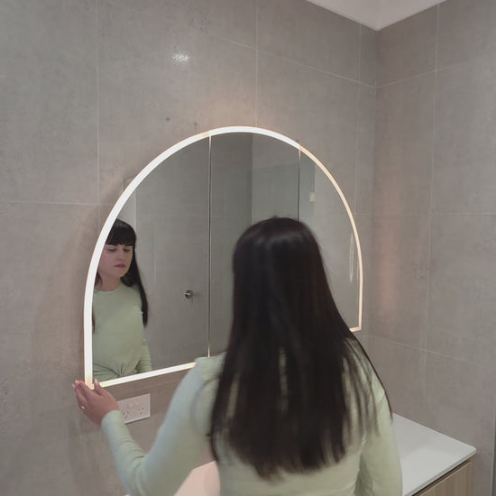 Female opening the door of an LED arched mirror cabinet showing adjustable glass internal shelves