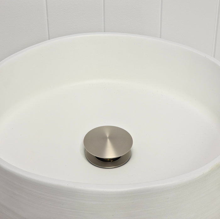 Lisbon Pop Up Basin Waste in Brushed Nickel, plug raised open in a round white basin by a white vertical-panelled wall