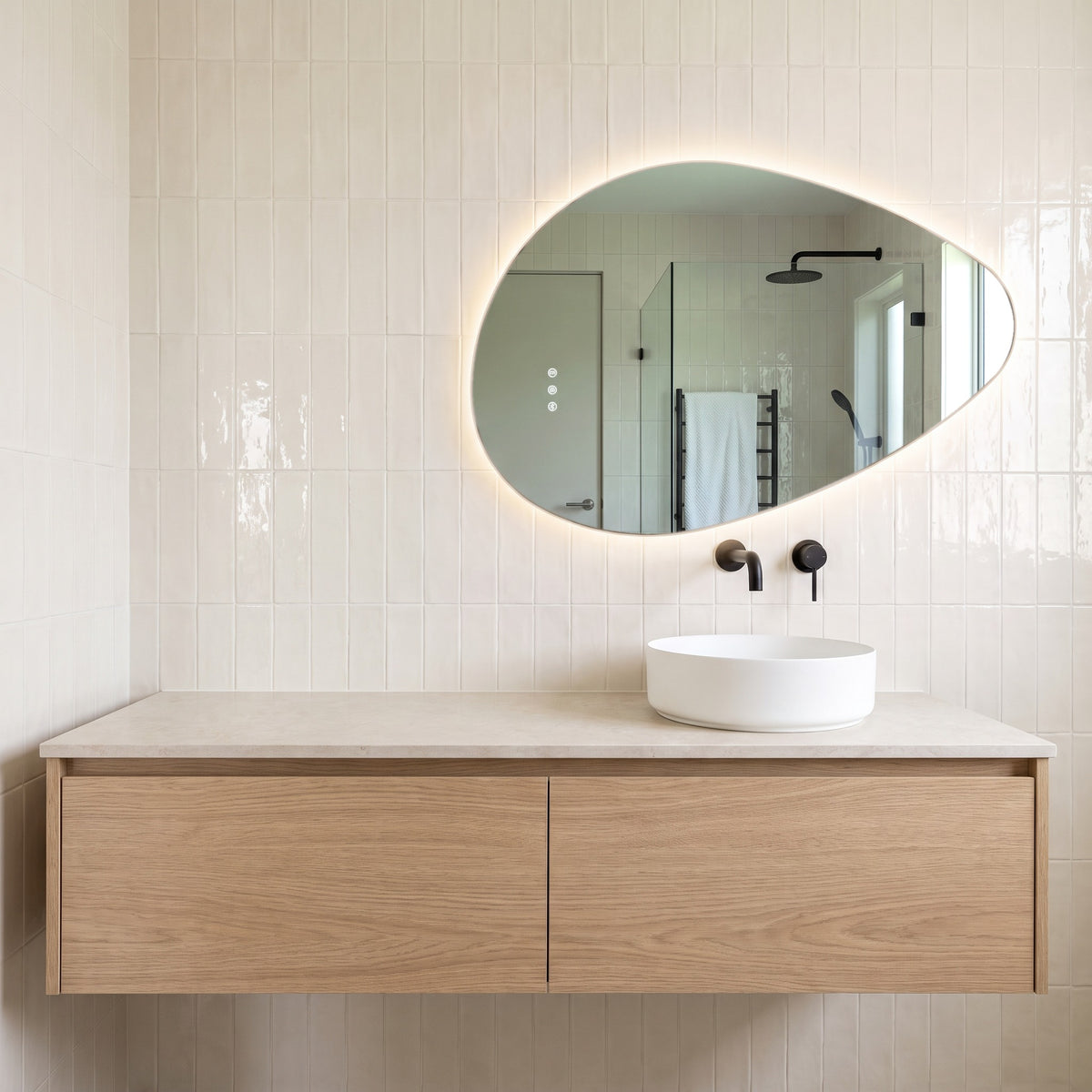 The Sorrento 800 x 550 mm pebble-shaped frameless backlit LED mirror, lit and hung landscape on white handmade tiles above a round white basin, matte black wall spout and mixer and an oak vanity