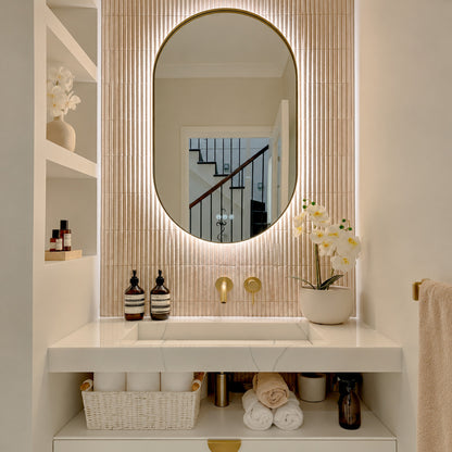 The Valencia 600 x 1000 mm oval framed backlit LED mirror in Brushed Brass, lit and seen straight-on above a white stone basin with a matching wall spout and mixer