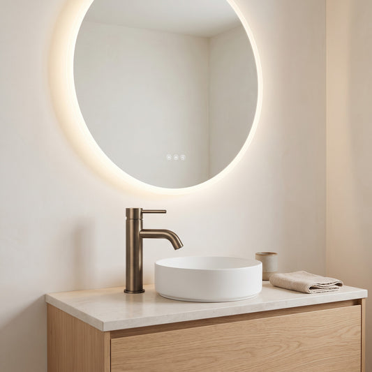 Vienna Short Basin Mixer in Antique Brass, beside a white vessel basin on oak below a backlit round mirror