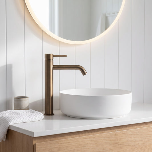 Vienna Tall Basin Mixer in Antique Brass, beside a white vessel basin on an oak vanity below a backlit round mirror