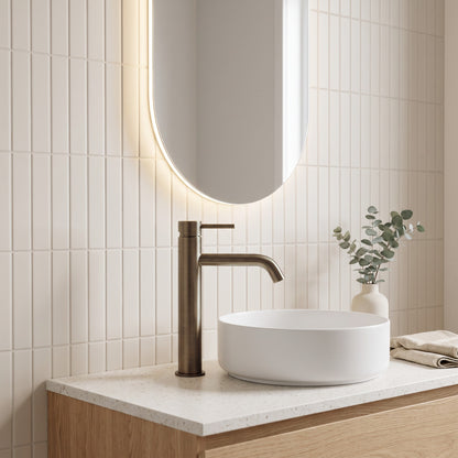 Vienna Tall Basin Mixer in Antique Brass, beside a white vessel basin on terrazzo below a backlit pill mirror